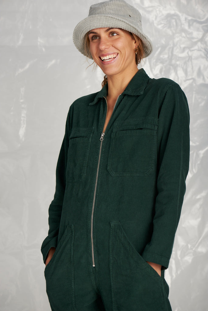 Femme debout de face en combinaison Velour verte avec poches. Elle porte un bob gris en Velour de la marque Graine