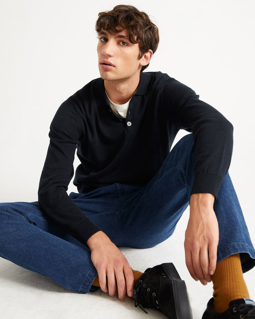 Un homme assis porte un jean et un pull avec col polo bleu marine de la marque Thinking Mu.