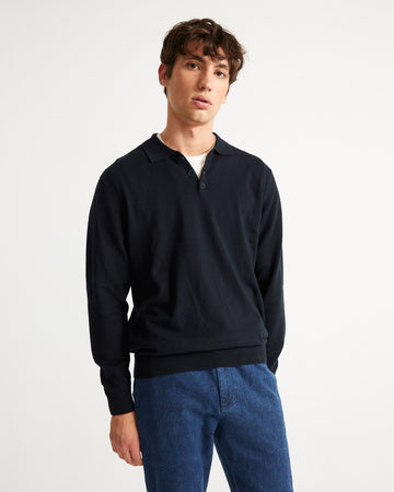Un homme debout porte un jean et un pull avec col polo bleu marine de la marque Thinking Mu.