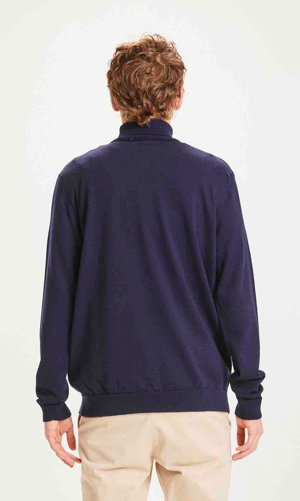 Un homme de dos porte un pull col roulé marine de la marque Knowledge Cotton Apparel.