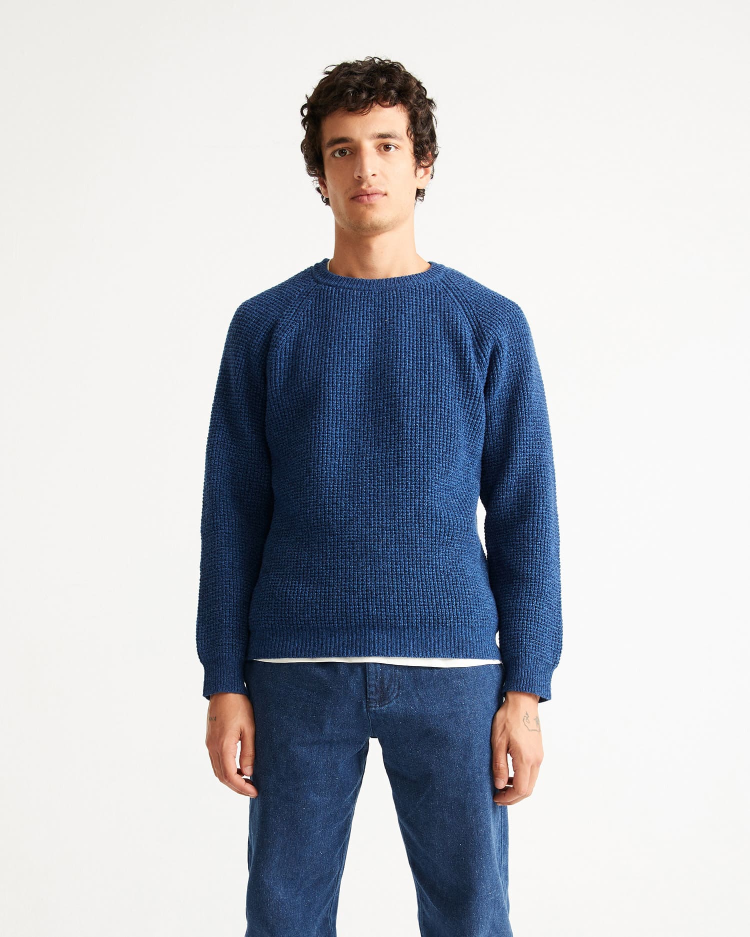 Un homme vue des genoux à la tête qui porte un jean et un pull en laine bleu de la marque Thinking Mu.